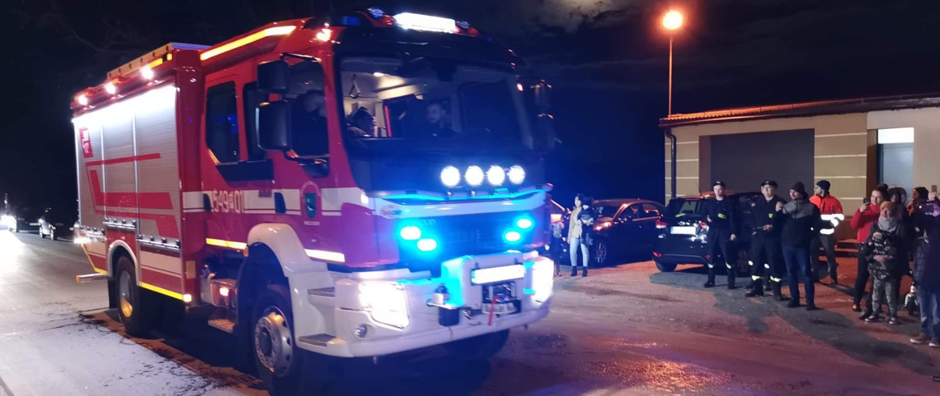 Na pierwszym planie widoczny wóz ratowniczo-gaśniczy przed strażnicą Ochotniczej Straży Pożarnej. Zdjęcie wykonane w porze nocnej na ulicy,
