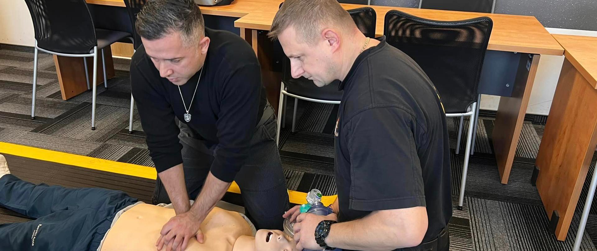 Na pierwszym zdjęciu strażacy-ratownicy wykonują symulację resuscytacji krążeniowo-oddechowej u osoby dorosłej. Jeden z ratowników wykonuje 30 uciśnięć klatki piersiowej, natomiast drugi wykonuje 2 oddechy ratownicze za pomocą worka samorozprężalnego.