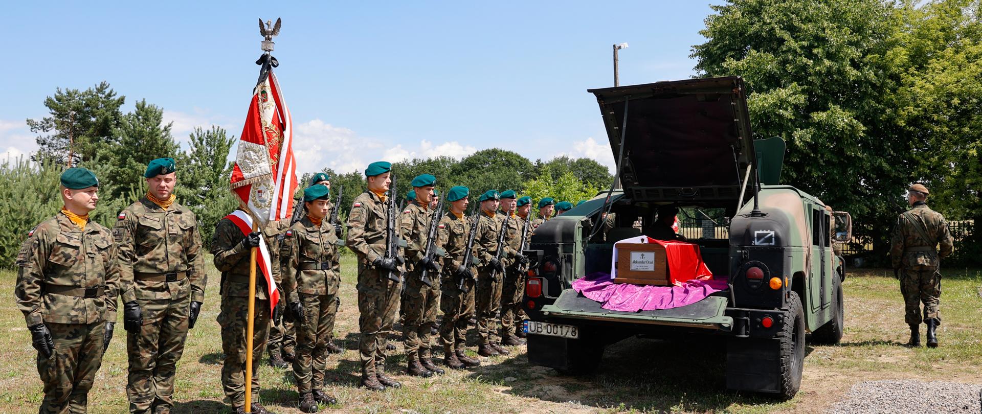 Żołnierze stoją w szyku. Na pierwszym planie trumna w samochodzie wojskowym.