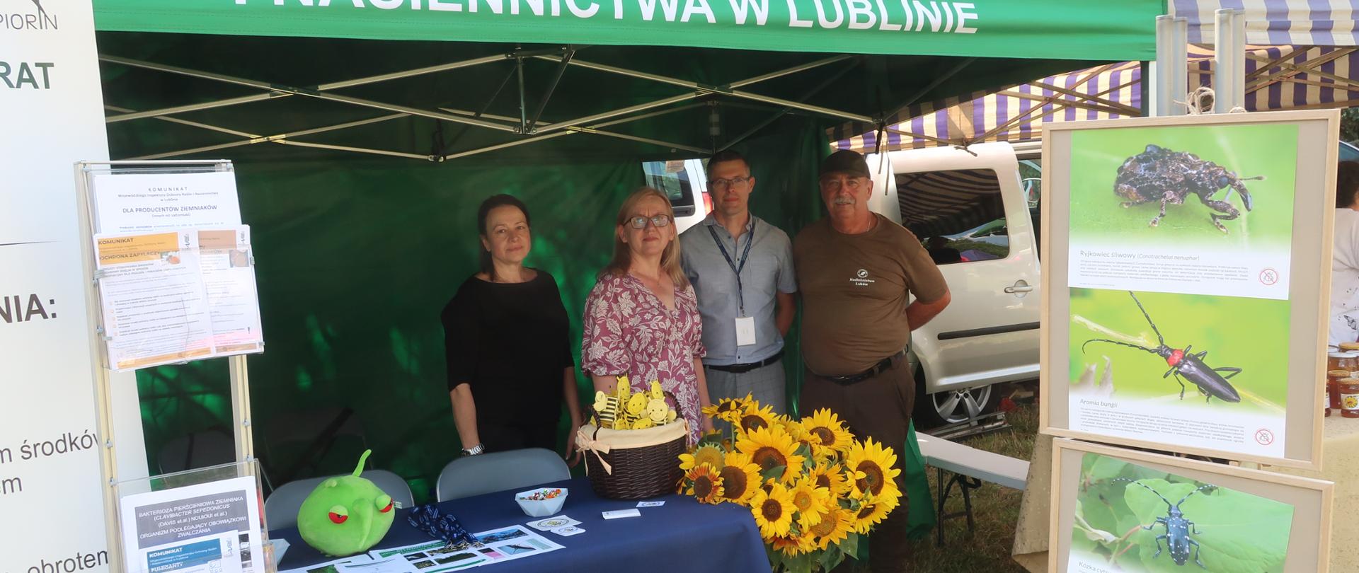 Dwie kobiety i dwóch mężczyzn stoi pod namiotem. Przed nimi ustawiony stolik z ulotkami i zieloną maskotką oraz kwiaty. Obok stojak z wystawą szkodników 
