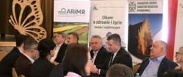 Przy stole siedzi grupa osób, która z uwagą słucha wygłaszanych prelekcji. Za nimi znajdują się roll-upy ARiMR, KRUS, PODR
