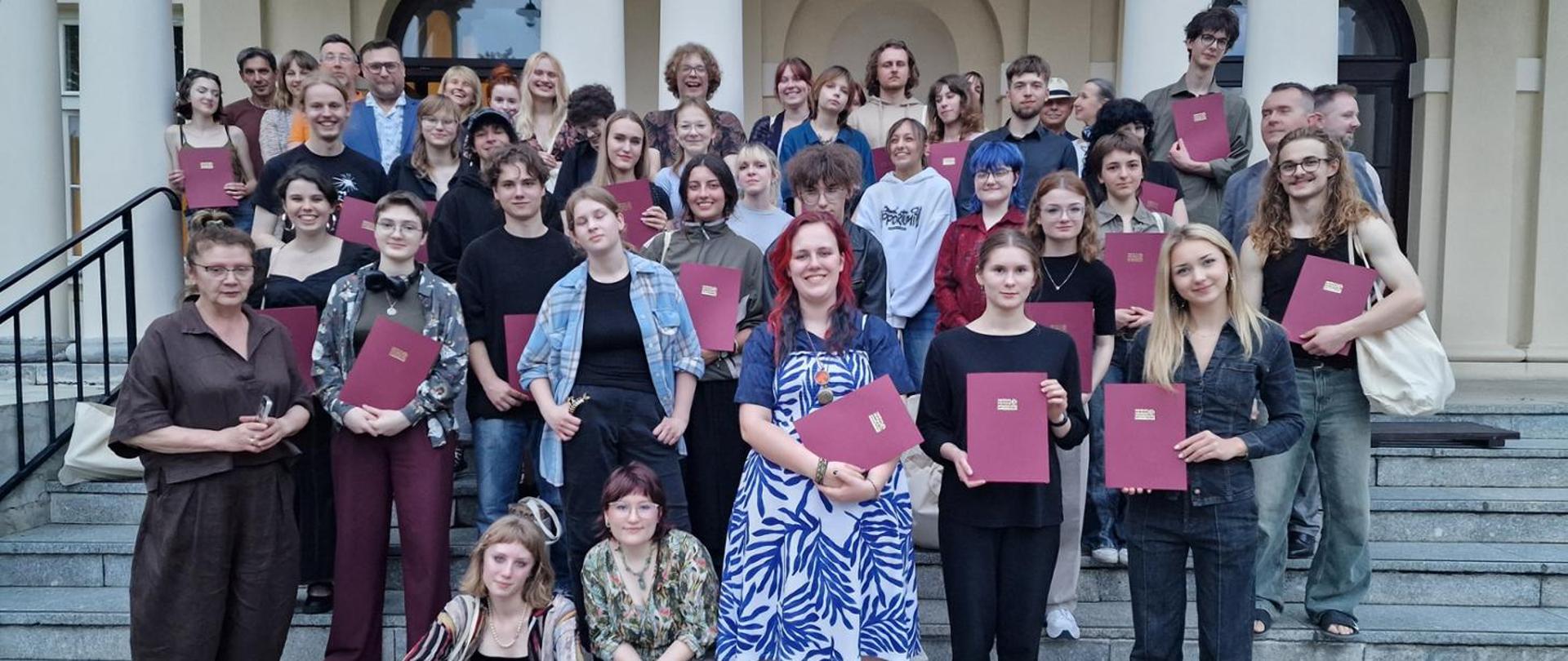 Na zdjęciu widzimy grupę Laureatów III etap Ogólnopolskiego Konkursu Plastycznego Centrum Edukacji Artystycznej Dłużew 2025 oraz Jurt Konkursu składające się z wykładowców uczelni artystycznych. Fotografia został wykonana na schodach domu plenerowego w Dłużewie. 