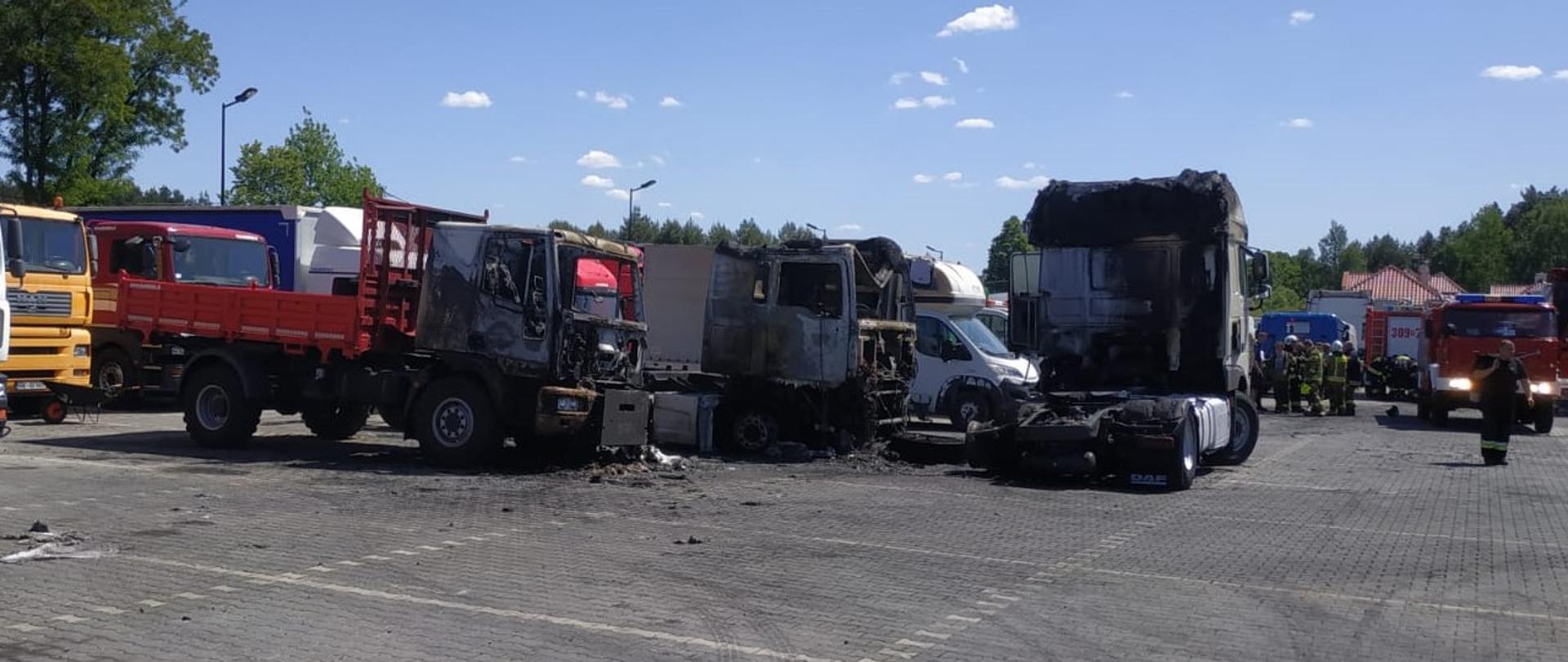 Zdjęcie przedstawia plac na terenie komisu samochodowego oraz trzy samochody ciężarowe, które uległy spaleniu. Po prawej stronie, w głębi widać samochód pożarniczy, a z lewej zaparkowane inne pojazdy.