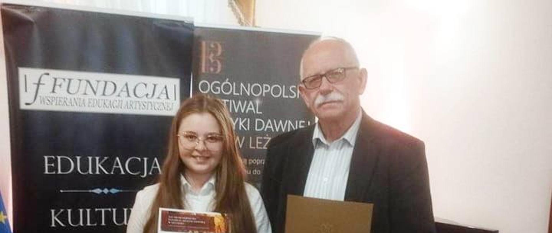 Dziewczynka trzymająca dyplom i czerwoną torbę, obok jej nauczyciel trzymający brązową teczkę,w tle baner XVI Ogólnopolskiego Festiwalu Muzyki Dawnej - Leżajsk 2023