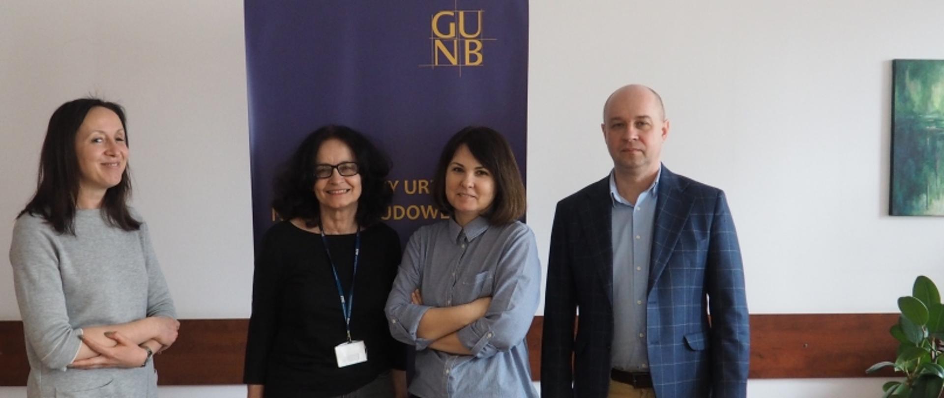 Cztery osoby stojące przed banerem z napisem „GUNB” i częściowo widocznym tekstem „Y UR DOW”.