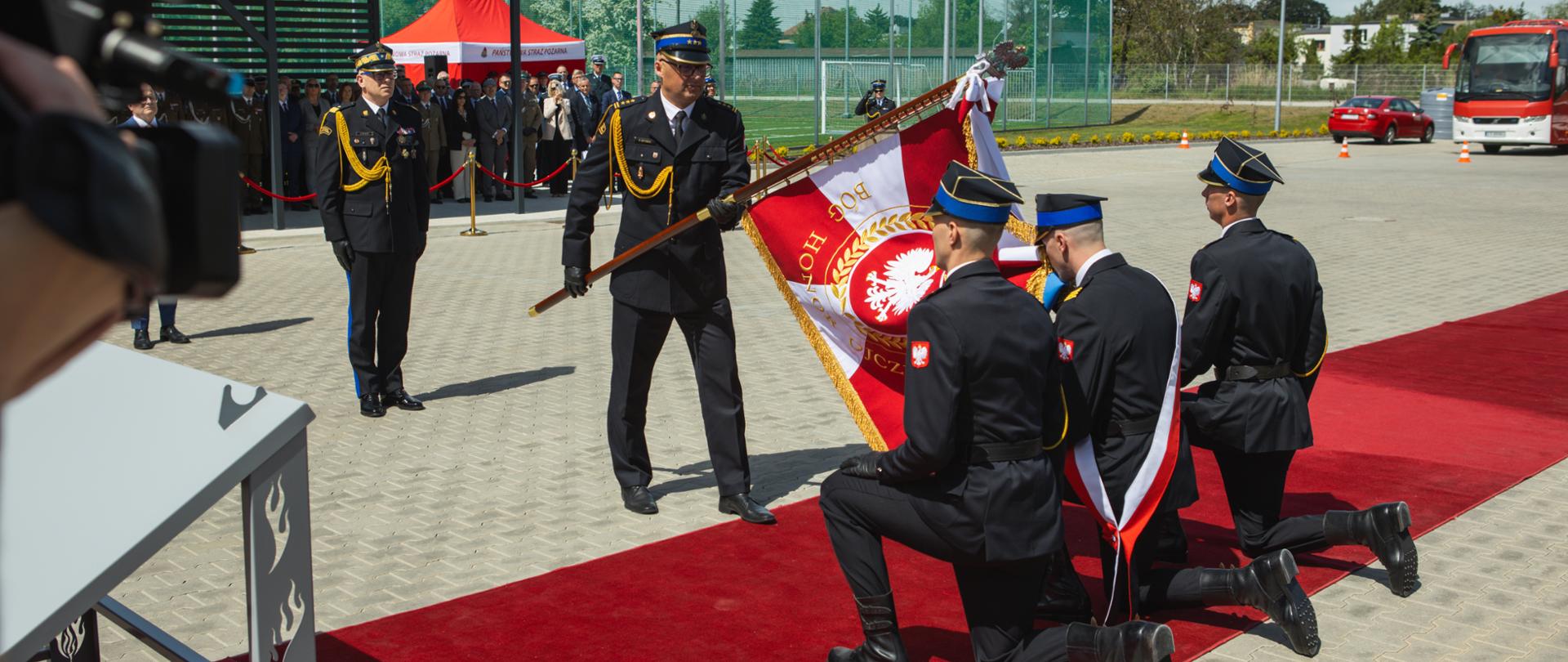 Strażacy pocztu sztandarowego klęczą na czerwonym dywanie podczas ceremonii przekazania sztandaru. Komendant miejski PSP w Grudziądzu prezentuje sztandar przed uczestnikami uroczystości.