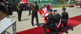 Strażacy pocztu sztandarowego klęczą na czerwonym dywanie podczas ceremonii przekazania sztandaru. Komendant miejski PSP w Grudziądzu prezentuje sztandar przed uczestnikami uroczystości.