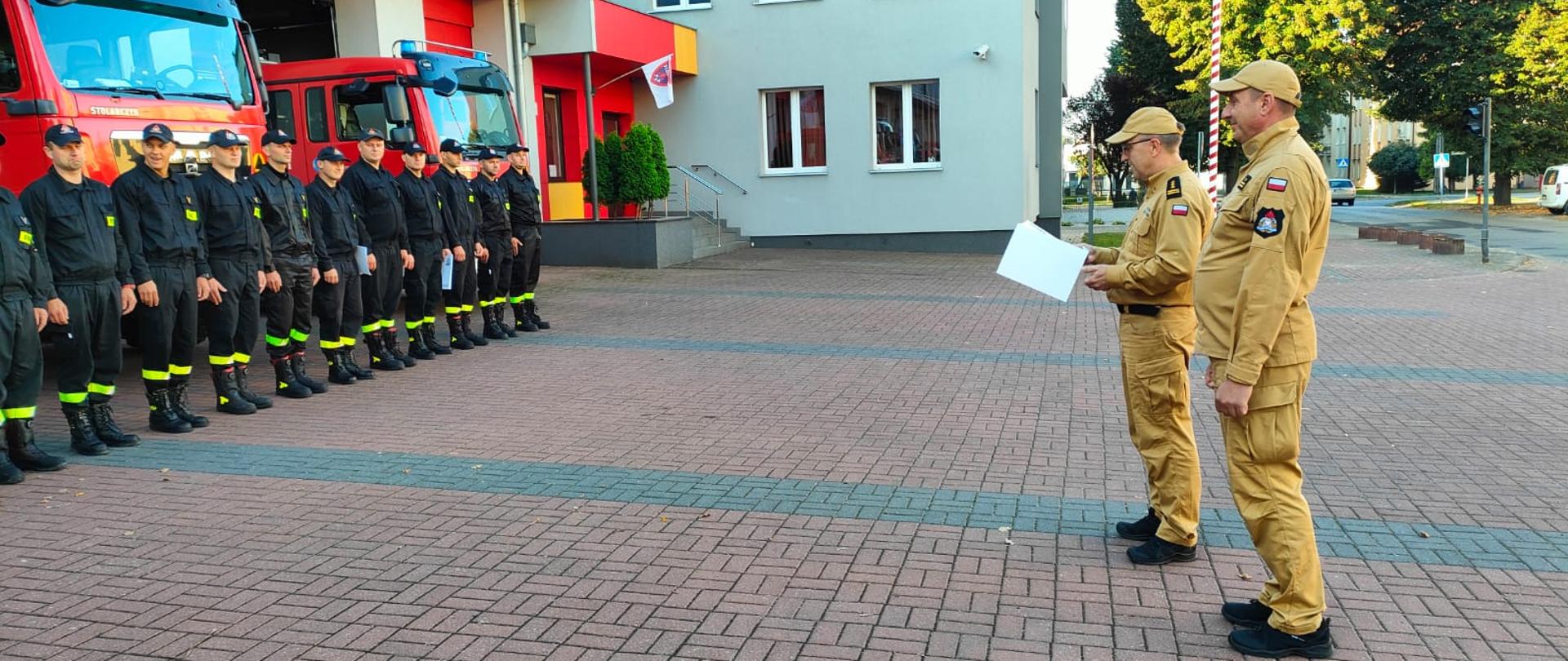 Mężczyzna w piaskowym mundurze odczytuje gratulacje przed grupą strażaków
