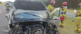 Kolizja na drodze ekspresowej. Jezdnia w kierunku Poznania. Na lewym pasie ruchu stoi samochód osobowy, który ma bardzo mocno uszkodzony przód. Auto ma uniesioną pokrywę silnika i otwarte drzwi od strony kierowcy, Przed samochodem na asfalcie stoi akumulator. Obok auta stoi 3 strażaków w czerwonych hełmach.
