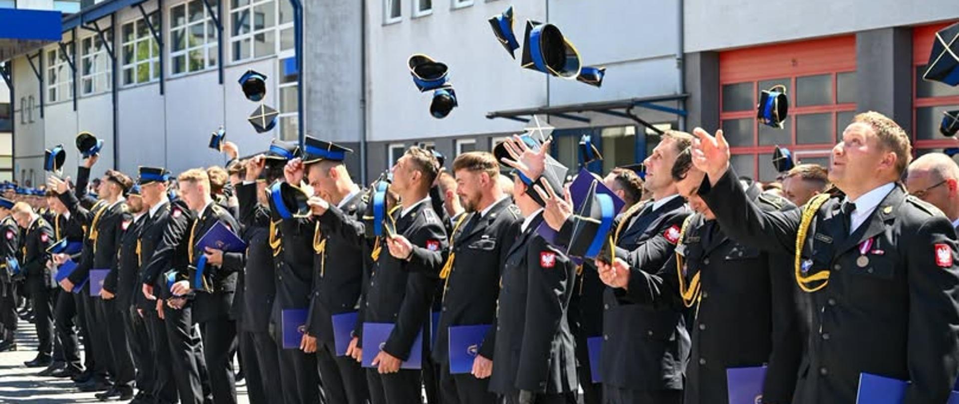 Zdjęcie przedstawia liczną grupę funkcjonariuszy Państwowej Straży Pożarnej podczas oficjalnej uroczystości. Strażacy stoją w szeregu na zewnątrz, przed nowoczesnym, jasnoszarym budynkiem z czerwonymi bramami garażowymi. Wszyscy ubrani są w ciemne mundury galowe ze złotymi akcentami (sznurami galowymi) i emblematami na ramionach.Wielu z nich patrzy w niebo, radosnym gestem podrzucając w górę tradycyjne czapki rogatywki z granatowymi otokami. W rękach trzymają niebieskie teczki, prawdopodobnie z aktami mianowania lub dyplomami. Atmosfera jest podniosła i radosna. W prawym dolnym rogu widnieje podpis autora zdjęcia: st. kpt. Piotr Krzak | Państwowa Straż Pożarna.