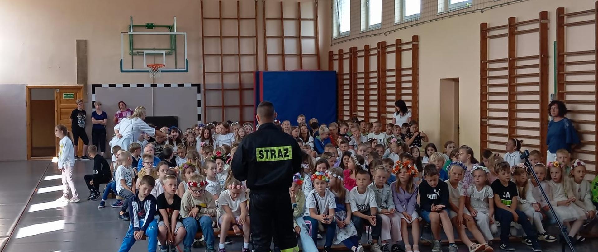 Dzieci w wieku szkolnym zgromadzone na sali sportowej siedzą na ławkach i słuchają strażaka ubranego w mundur koszarowy.