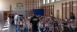 Dzieci w wieku szkolnym zgromadzone na sali sportowej siedzą na ławkach i słuchają strażaka ubranego w mundur koszarowy.