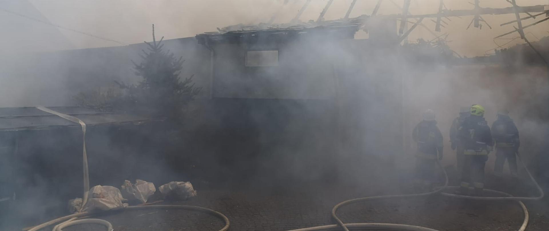 Zdjęcie przedstawia strażaków podczas działań gaśniczych przy pożarze zabudowań gospodarczych w miejscowości Łany.
Na pierwszym planie widoczne odcinki strażackie napełnione wodą. W oddali w dymie widoczny spalony budynek i spaloną więźbę dachową.