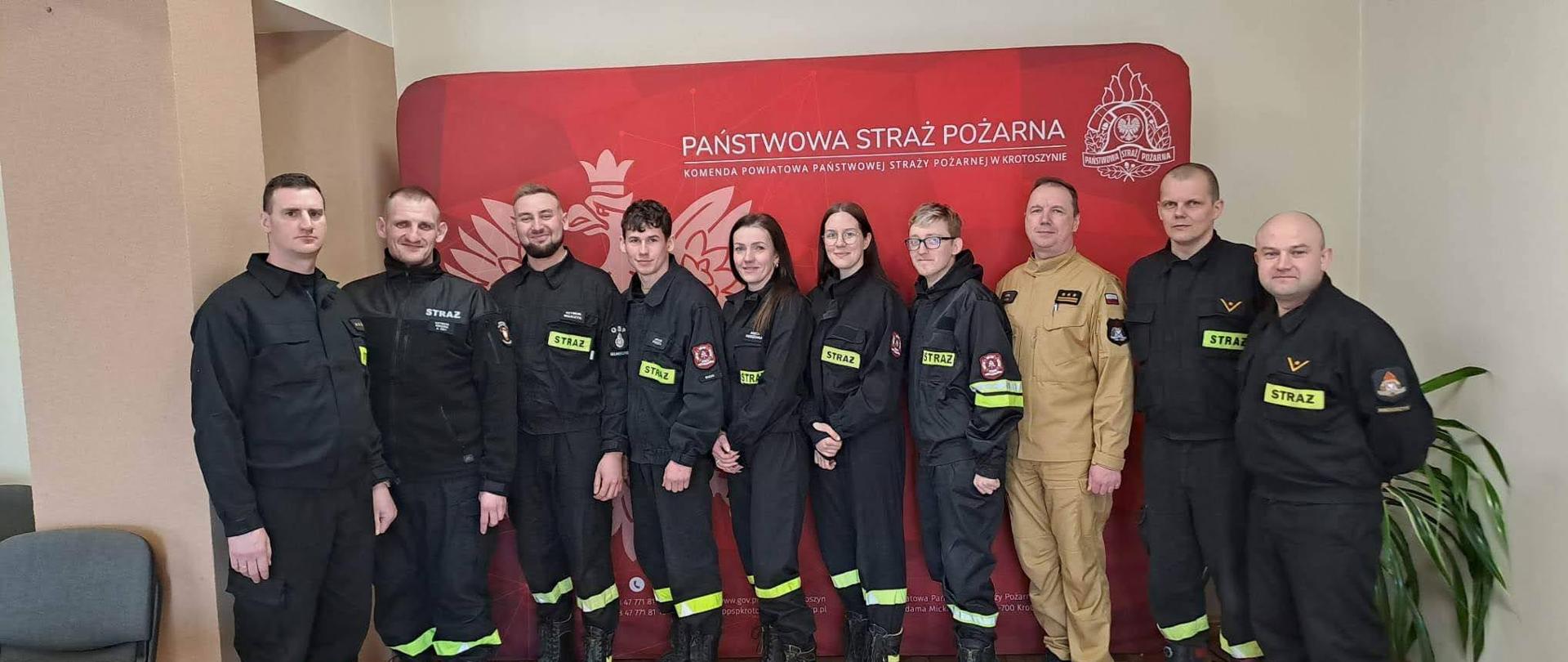 Zdjęcie grupowe na tle baneru komendy powiatowej absolwentów szkolenia z zakresu kwalifikowanej pierwszej pomocy z Komendantem Powiatowym PSP w Krotoszynie oraz członkami komisji egzaminacyjnej.