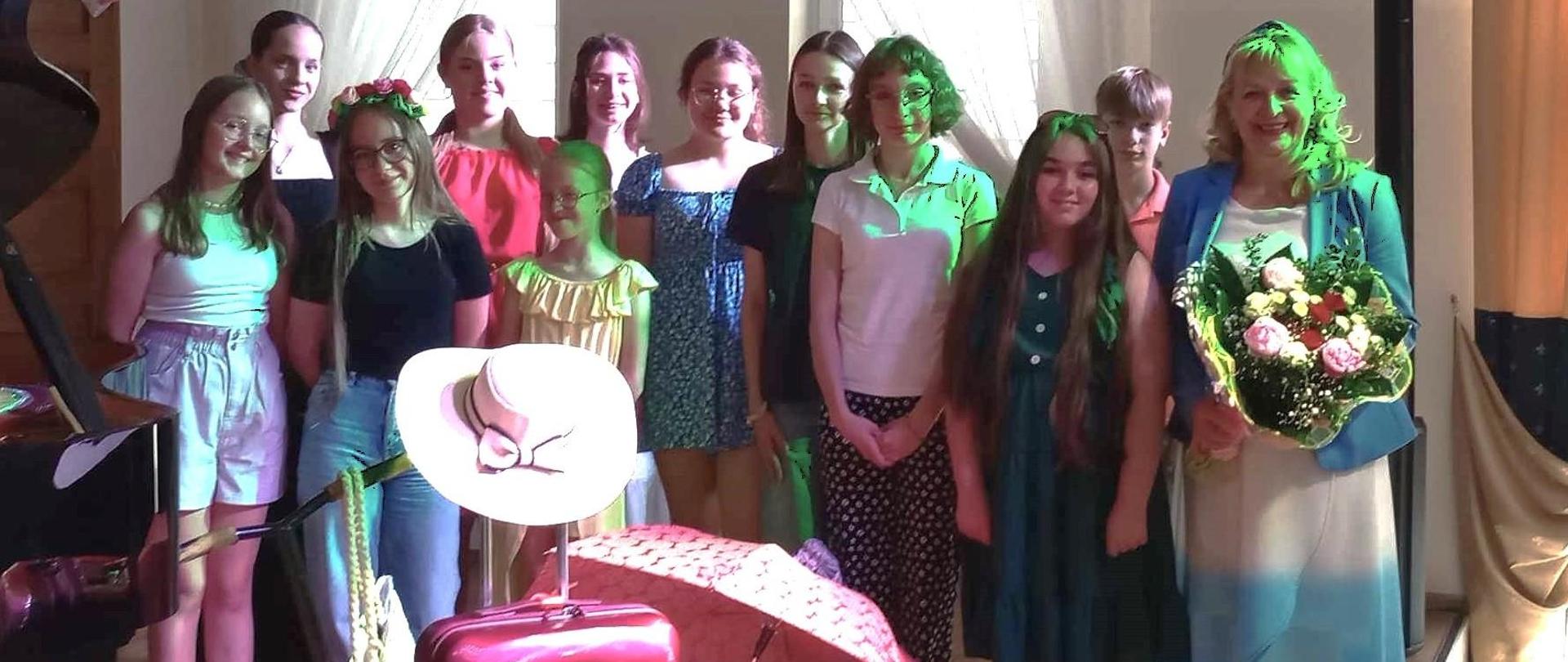 Zdjęcie zbiorowe na scenie przy fortepianie. Dziewczęta i chłopiec kolorowo ubrani stoją ustawieni w dwóch rzędach. Przed nimi walizki, klapki plażowe, piłka, hulajnoga. Nauczycielka trzyma w ręku kwiaty.