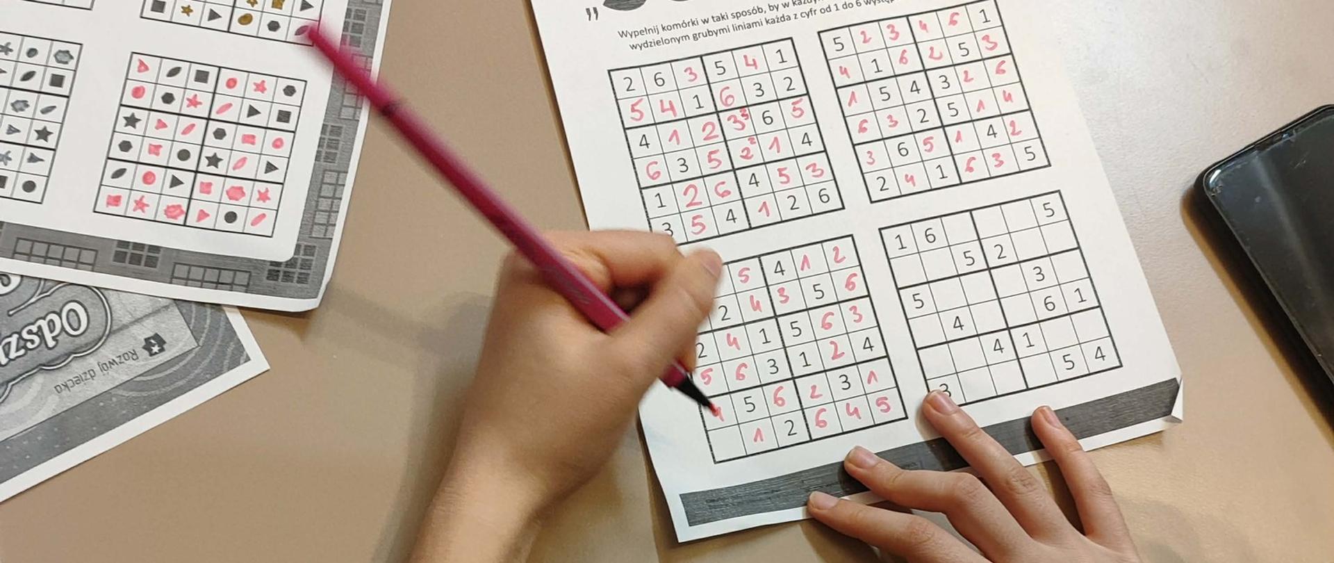 Na zdjęciu widać ręce dziewczynki trzymającej ołówek nad kartką sudoku