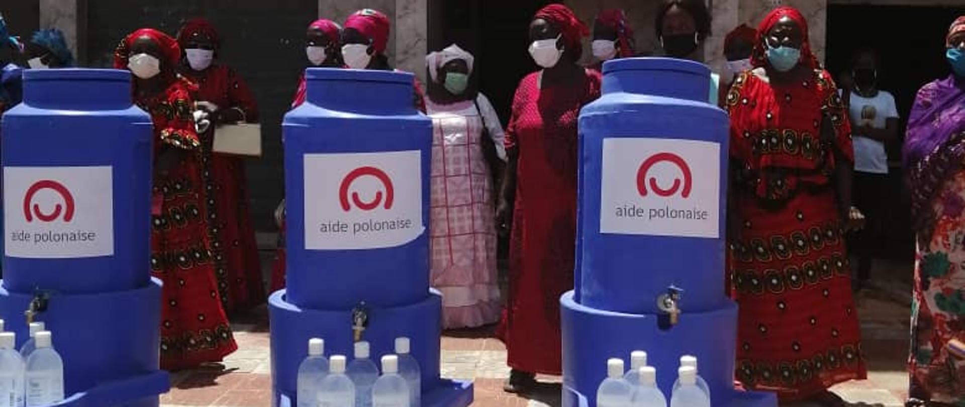 na podłodze stoją trzy pojemniki na wodę, a w tle stoją kobiety ubrane w kolorowe ubrania