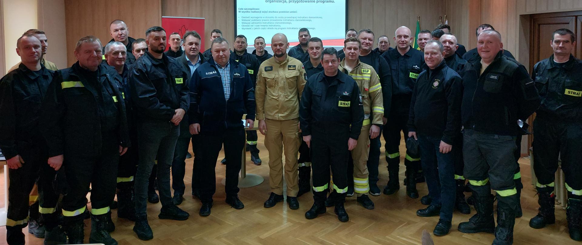 Strażacy druhowie Ochotniczych Straży Pożarnych stoją w sali wraz ze strażakami państwowej straży pożarnej, w tle wisi ekran multimedialny