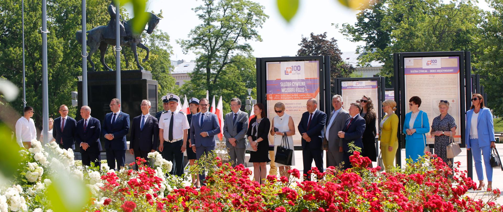 Na zdjęciu znajdują się elegancko ubrani urzędnicy - około 20 osób, panowie w garniturach, panie w żakietach. Na pierwszym planie znajdują się krzewy czerwonych i białych róż. Za ludźmi znajdują się plansze z plakatami. Widać, że jest słoneczny dzień, kilka osób ma założone okulary przeciwsłoneczne.