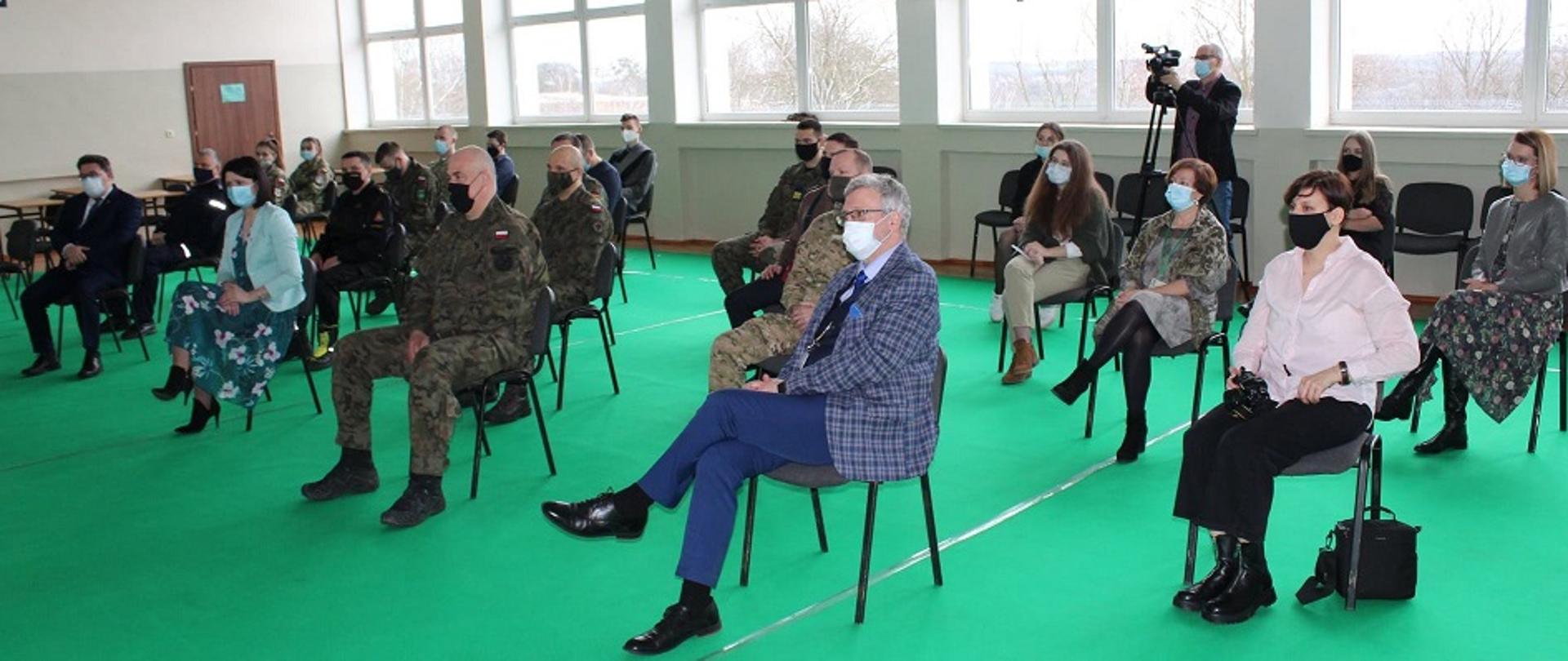NA ZDJĘCIU WIDAĆ OSOBY Z MASECZKAMI NA TWARZACH SIEDZĄCE NA KRZESŁACH, ZA NIMI STOI MĘŻCZYZNA TRZYMA W DŁONI KAMERĘ OSADZONĄ NA STATYWIE.