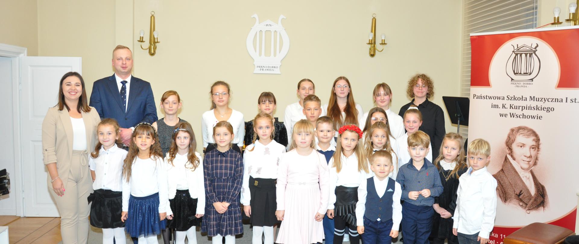 Dwudziestu trzech uczniów, z lewej strony dwie osoby dorosłe. Z prawej strony baner z nazwą i logiem szkoły oraz szkicem patrona szkoły. W tle na ścianie widoczne kinkiety oraz logo szkoły.