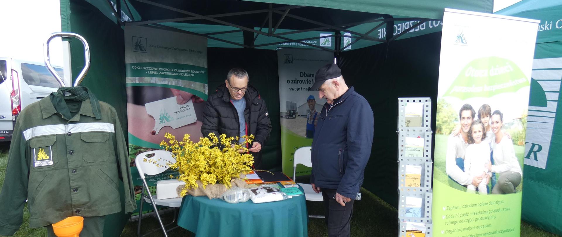 Stoisko Placówki Terenowej KRUS w Wysokiem Mazowieckiem pod namiotem z logo KRUS po lewej stronie. Wewnątrz namiotu pracownik Kasy zapoznaje mężczyznę z materiałami informacyjnymi KRUS. Z lewej strony namiotu znajduję się wzorcowa odzież robocza oraz wał odbioru mocy, z prawej strony zielony baner Kasy oraz ulotki.