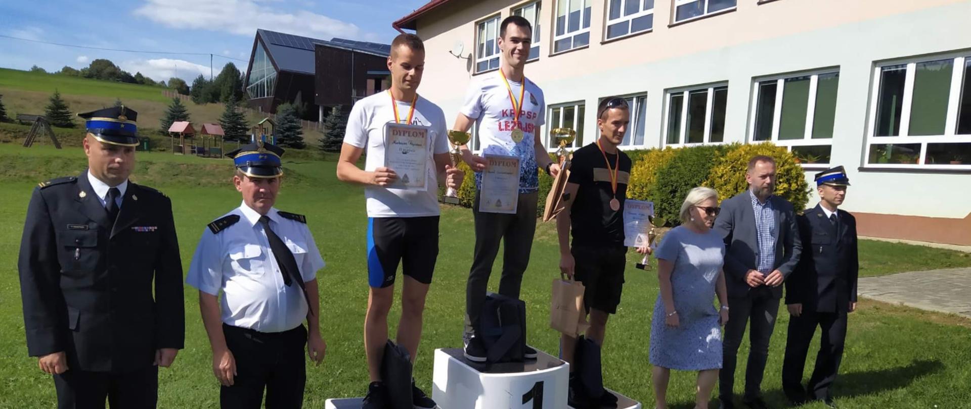 Na zdjęciu widzimy strażaków w ubraniach sportowych, zwycięzców biegu, stojących na podeście z medalami na szyi oraz dyplomami i pucharami w rękach, obok stoją osoby w ubraniach cywilnych oraz mundurach galowych. W tle widzimy część budynku oraz plac zabaw.