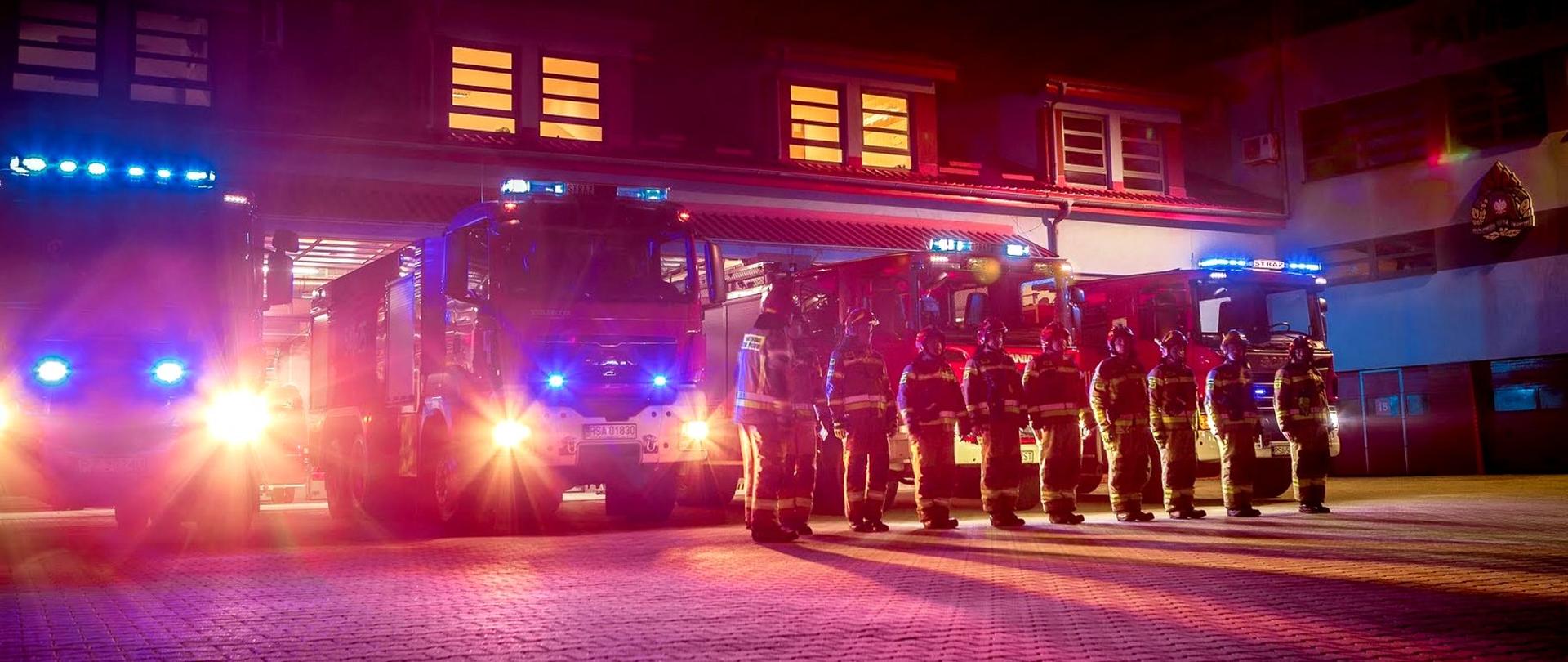 Zdjęcie zrobione przed jednostką ratowniczo-gaśniczą. Na fotografii widać cztery samochody gaśnicze z włączonymi sygnałami świetlnymi. Przed samochodami, w szeregu stoją strażacy ubrani w mundury specjalne.