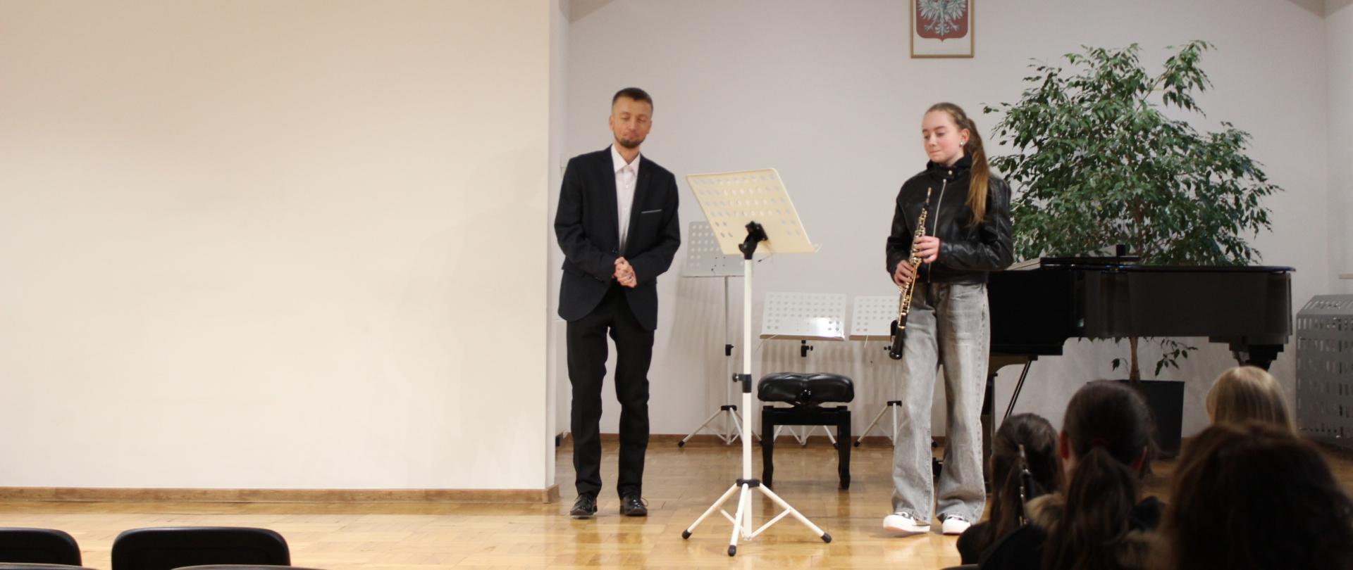 na scenie sali koncertowej stoją nauczyciel oraz uczennica z obojem w dłoniach, w tle głowy publiczności