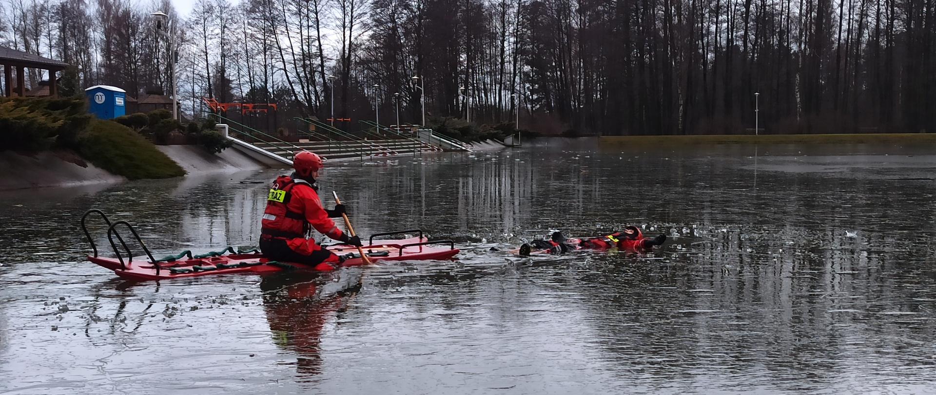 Na zdjęciu widać dwóch strażaków ubranych w ubrania do ratownictwa wodno-lodowego. Jeden z strażaków znajduje się w wodzie udając osobę poszkodowaną, drugi zaś dopływa do niego na saniach lodowych. W tle infrastruktura OSIR "Gutwin"
