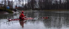 Na zdjęciu widać dwóch strażaków ubranych w ubrania do ratownictwa wodno-lodowego. Jeden z strażaków znajduje się w wodzie udając osobę poszkodowaną, drugi zaś dopływa do niego na saniach lodowych. W tle infrastruktura OSIR "Gutwin"