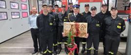 Strażacy wraz z kończącym służbę Jackiem Dąbkiem stoją na tle drabiny pożarniczej. Jacek trzyma pamiątkową tablicę, przed nim kosz z kwiatami.