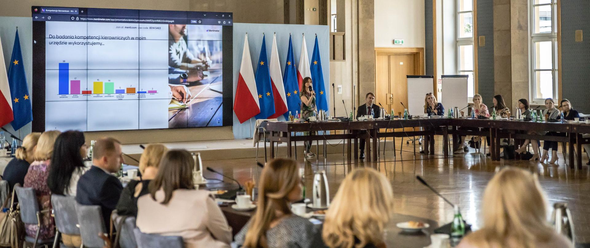 Kobieta stoi i przemawia do mikrofonu, za nią widać ekran na którym wyświetlana jest prezentacja. Obok ekranu flagi Polski i Unii Europejskiej