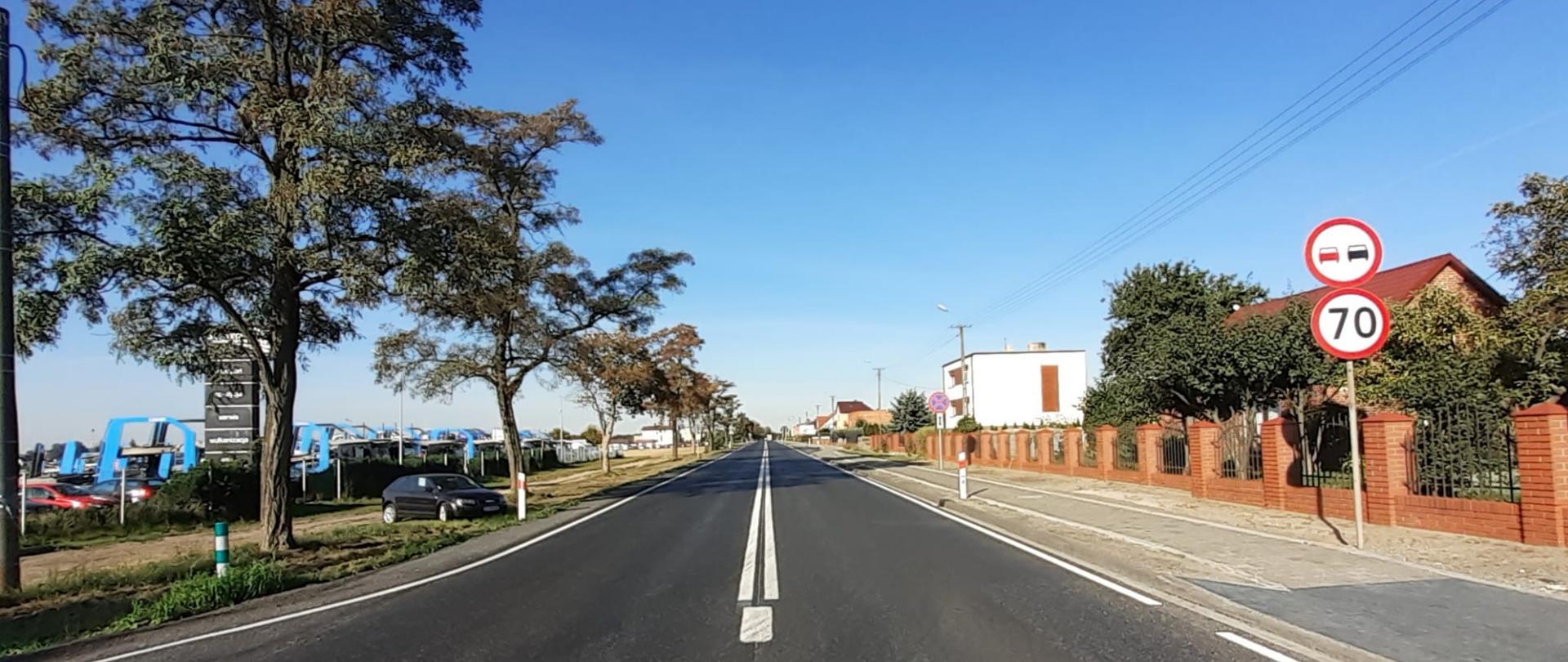 Zdjęcie przedstawia drogę krajową nr 1. Na zdjęciu widoczna droga, po jednym pasie w każdym kierunku. Z prawej strony chodnik. , znak ograniczenia prędkości do 70 kilometrów na godzinę oraz zakaz wyprzedzania. Zarówno po prawej stronie jak i lewej widoczne zabudowania. 