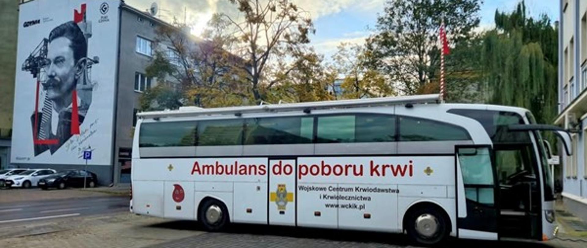 Biały ambulans oznaczony jako „Ambulans do poboru krwi”. Pojazd zaparkowany jest na ulicy, a w tle widać budynek z dużym muralem przedstawiający mężczyznę z wąsami Tadeusza Wendę.