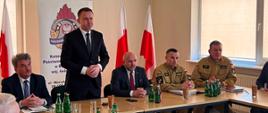Na zdjęciu zastępca świętokrzyskiego komendanta wojewódzkiego PSP oraz komendant powiatowy PSP w Busku-Zdroju w umundurowaniu służbowym, dwóch Posłów RP oraz Wicestarosta Powiatu buskiego przy stole prezydialnym. Jeden z Posłów zabiera głos. W tle flagi Polski oraz baner z logiem PSP.