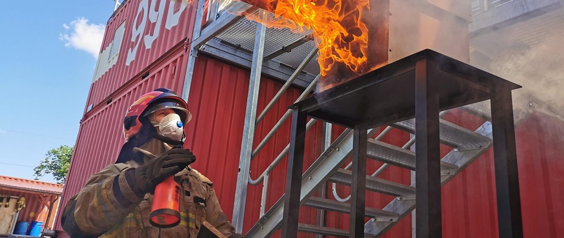 strażak w hełmie patrzy na ogień, w tle czerwone baraki