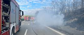 autostrada na której stoją pojazdy gaśnicze, po prawej stronie widoczny dym z płonącego pojazdu oraz wypalone od ognia pobocze drogi 