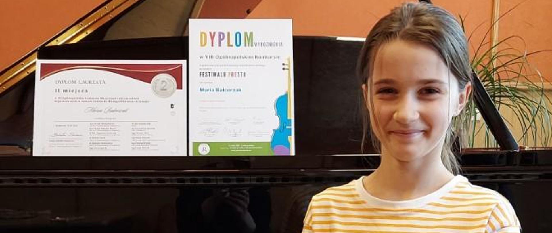 Zdjęcie kolorowe. Uśmiechnięta dziewczynka z ciemnymi włosami, ubrana w koszulkę w paski siedzi przed otwartym czarnym fortepianem. Na pulpicie instrumentu stoją dwa dyplomy z konkursów.