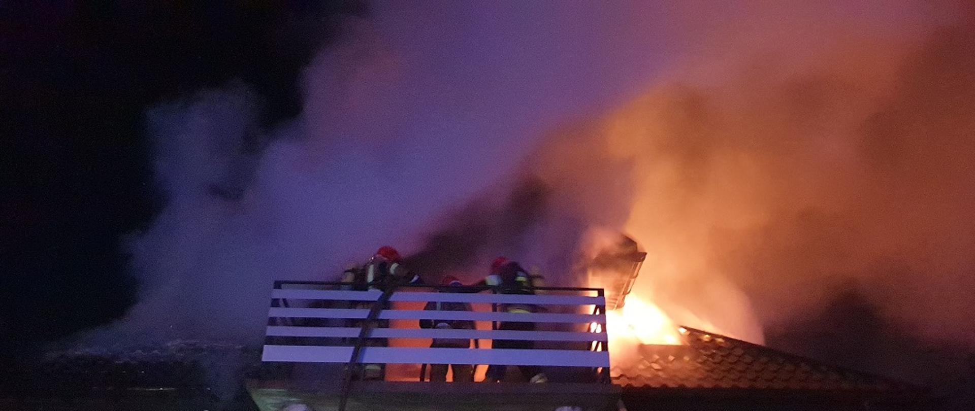 Na zdjęciu widzimy działania gaśnicze na palącym się poddaszu budynku mieszkalnego jednorodzinnego. Strażacy zabezpieczeni w aparaty ochrony dróg oddechowych gaszą palącą się konstrukcję dachu.