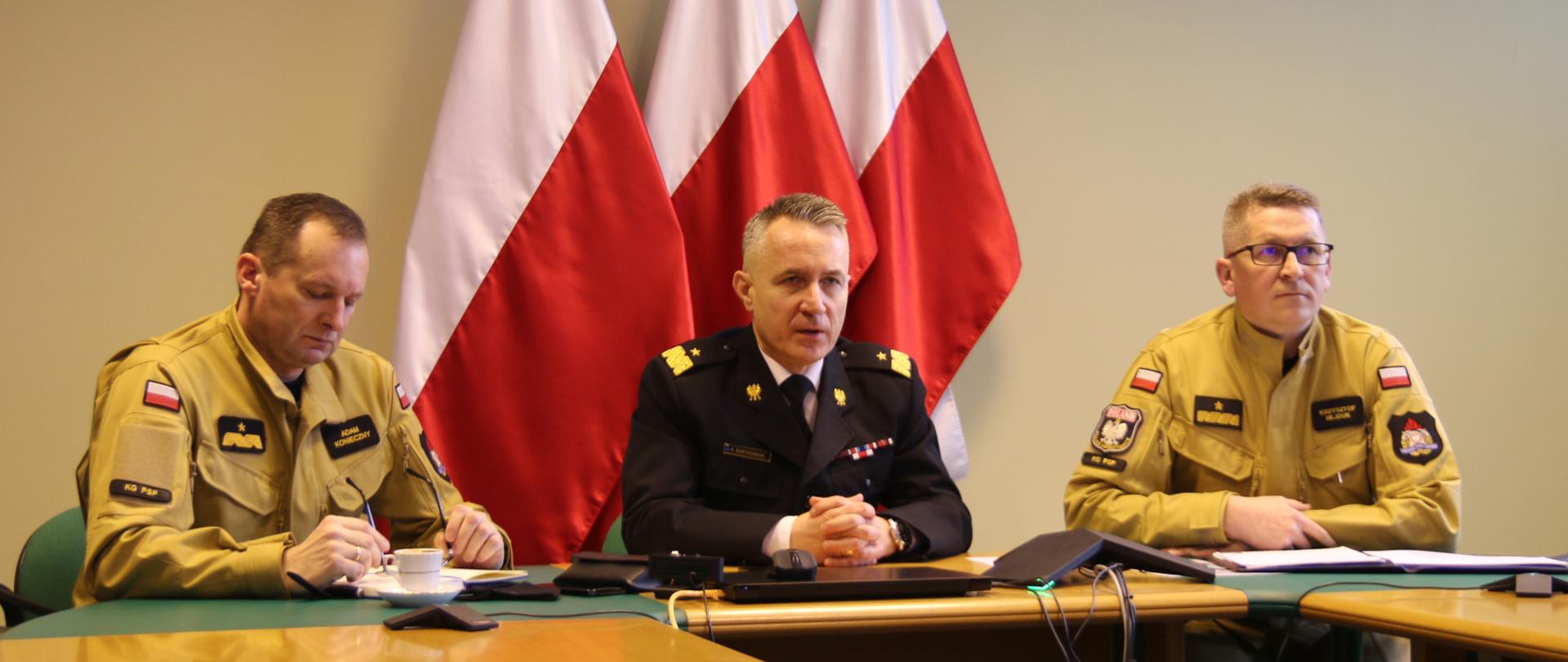 Kierownictwo Komendy Głównej Państwowej Straży Pożarnej podczas wideokonferencji. W tel flagi państwowe oraz wiszące na ścianie godło
