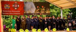 Pod zadaszeniem sceny ustawieni są przedstawiciele różnych służb mundurowych, samorządowcy oraz goście honorowi, w tym wojewoda, marszałek, parlamentarzyści i kierownictwo PSP. W tle baner obchodów z logotypami PSP, ZOSP RP oraz zdjęciem działań gaśniczych. Na podeście dekoracja ze zwiniętych węży i sprzętu pożarniczego.