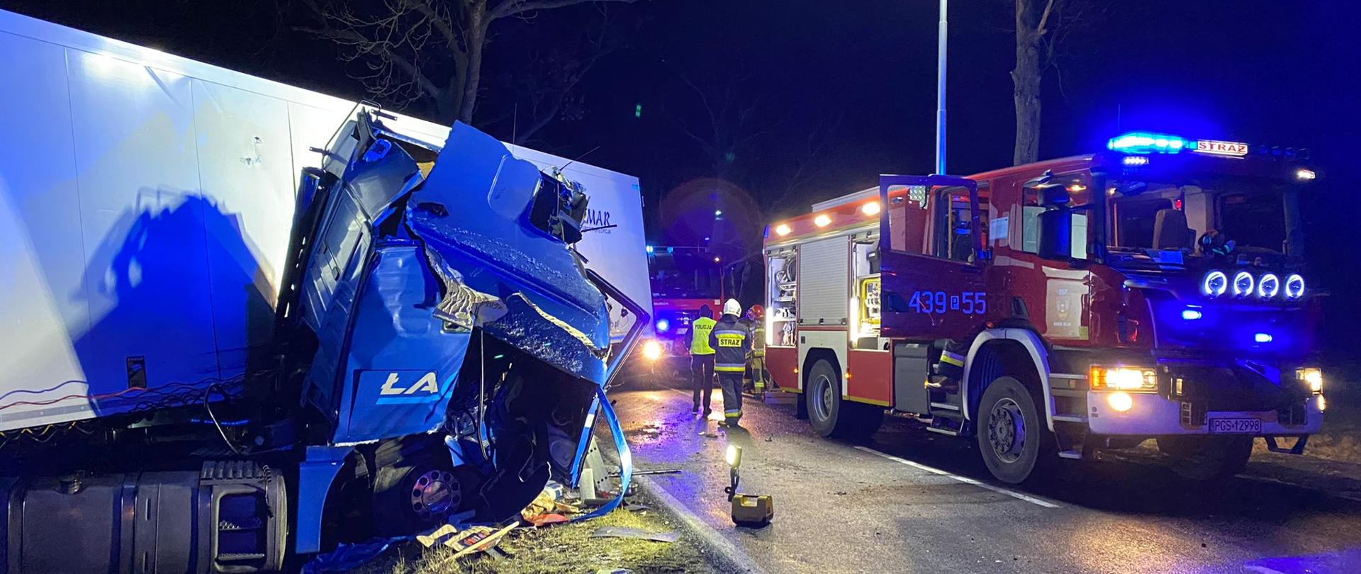 Na zdjęciu widać uszkodzoną ciężarówkę poza drogą oraz zastęp straży pożarnej. 