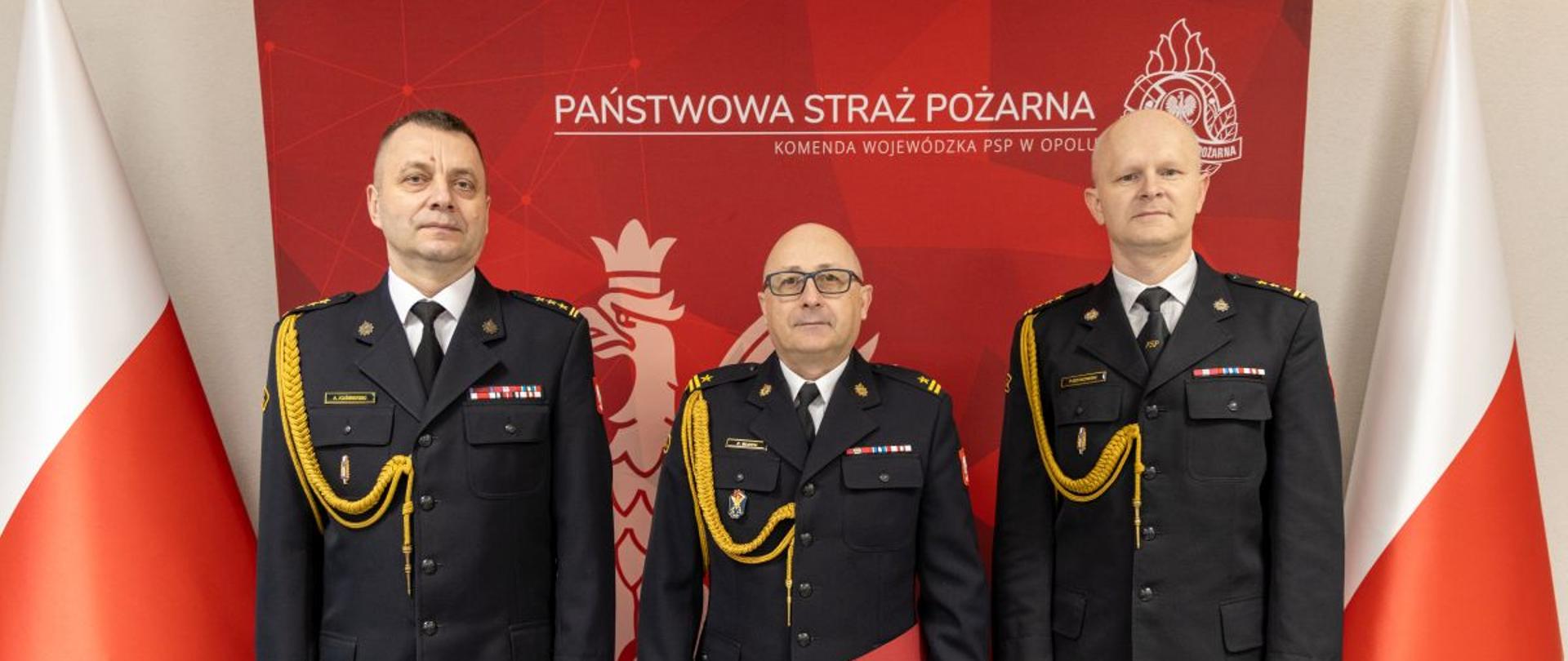 Mł. bryg. Zenon Błoch stoi pomiędzy st. bryg. Arkadiuszem Kuśmierskim i st. bryg. Pawłem Gotkowskim podczas uroczystości powierzenia obowiązków Zastępcy Komendanta Powiatowego PSP w Nysie. Wszyscy w umundurowaniu galowym, na tle czerwonej ścianki z godłem PSP i polskimi flagami.