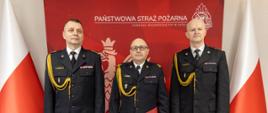 Mł. bryg. Zenon Błoch stoi pomiędzy st. bryg. Arkadiuszem Kuśmierskim i st. bryg. Pawłem Gotkowskim podczas uroczystości powierzenia obowiązków Zastępcy Komendanta Powiatowego PSP w Nysie. Wszyscy w umundurowaniu galowym, na tle czerwonej ścianki z godłem PSP i polskimi flagami.