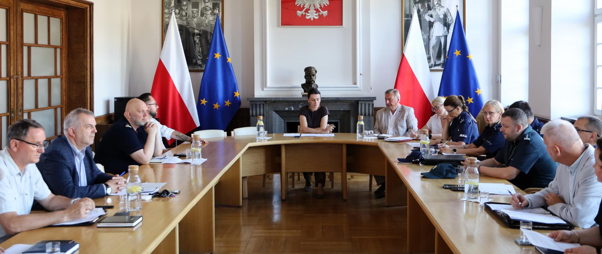 Sala konferencyjna z długim stołem, przy którym siedzą uczestnicy spotkania. Na środku, naprzeciwko wejścia, siedzi prowadząca obrady (wojewoda). W tle widoczne są dwie polskie flagi, dwie flagi Unii Europejskiej oraz godło Polski nad kominkiem. Po obu stronach wiszą czarno-białe fotografie historyczne, a uczestnicy mają przed sobą notatniki i dokumenty.