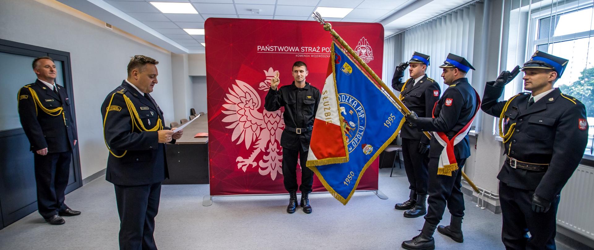 Ślubowanie str. Dominika Okonia. Ślubowanie składane przed Opolskim Komendantem Wojewódzkim PSP w Opolu st. bryg. Krzysztofem Kędrykiem oraz pocztem sztandarowym KW PSP w Opolu. W tle widoczny banner Państwowej Straży Pożarnej w kolorze czerwonym. W lewej stronie bryg. Ryszard Sałek naczelnik wydziału technicznego KW PSP w Opolu.