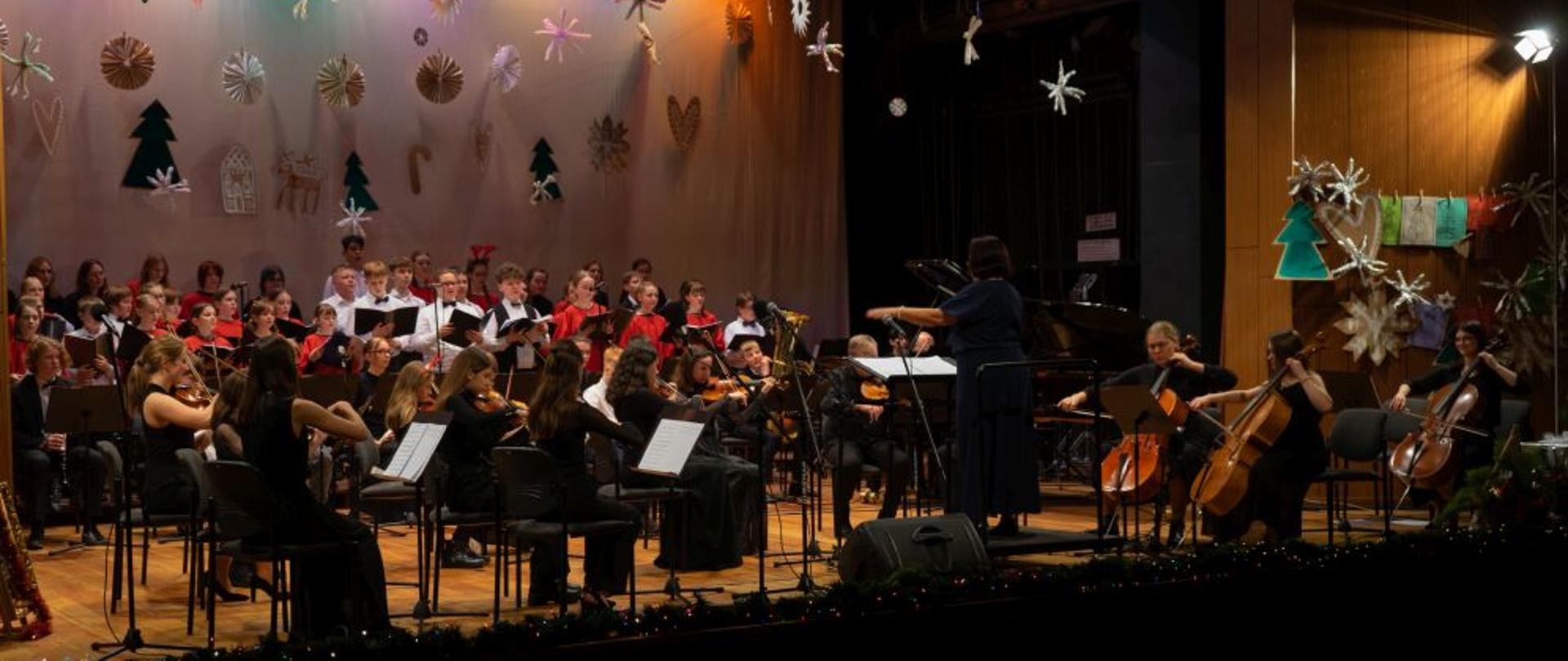 Zdjęcie kolorowe. Orkiestra (muzycy w czarnych strojach) i chór (w strojach czerwonych) na scenie udekorowanej elementami świątecznymi. Na pierwszym planie dyrygentka. 