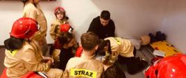 Na zdjęciu widzimy strażaka, który tłumaczy dzieciom uczestniczącym w zajęciach edukacyjnych zasady udzielania pierwszej pomocy.