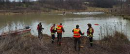 Strażacy ubrani w kamizelki ratunkowe stoją na brzegiem jeziora obok łódź aluminiowa na dalszym planie widać ćwiczących strażaków